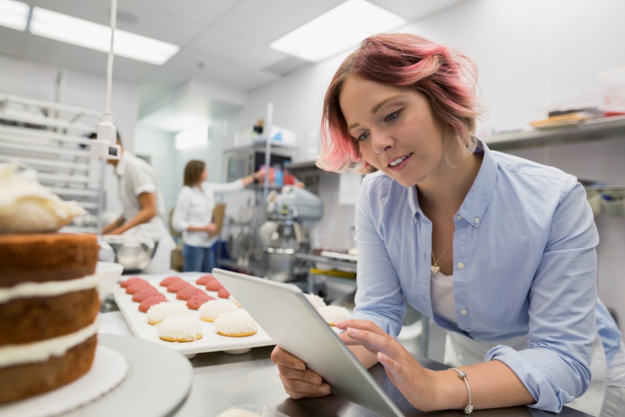 A baker in an industrial kitchen checks a tablet by a metal table with a decorated cake and sheet of cookies